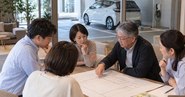駐車場の充電器が見える共用ラウンジで、配置図を囲んで話すマンション住民